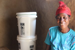 A woman, Catherine, sitting next to her SAM3 household filter which provides clean water for her family. She is smiling.